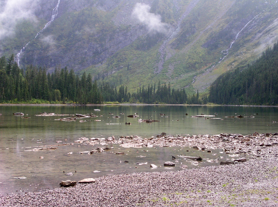 [Lots of stones near the shore and stones amid the lake as if the water levels may be lower than maximum. On the far shore are evergreens and the lower edge of what appears to be a large steep mountain.]